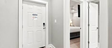 A hallway with a white door, a doormat, and a bed in the adjacent room.