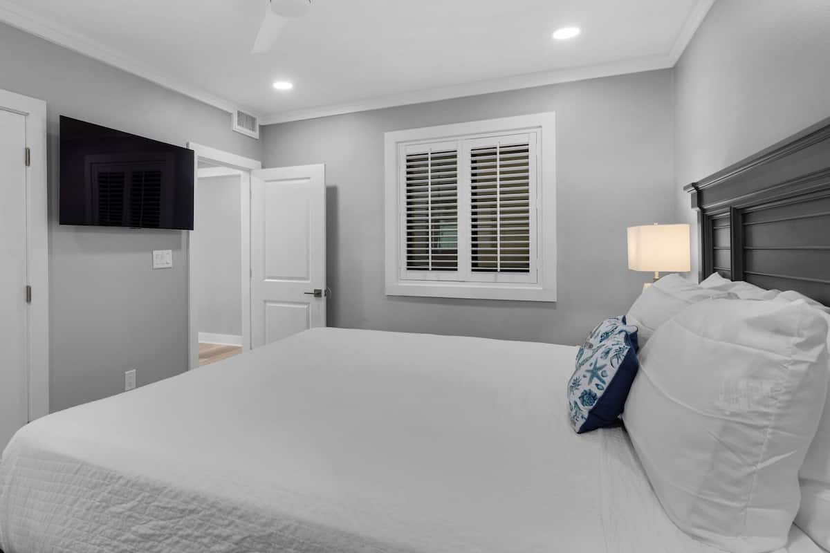 A bedroom with a large bed, a window with shutters, a TV mounted on the wall, and a door.