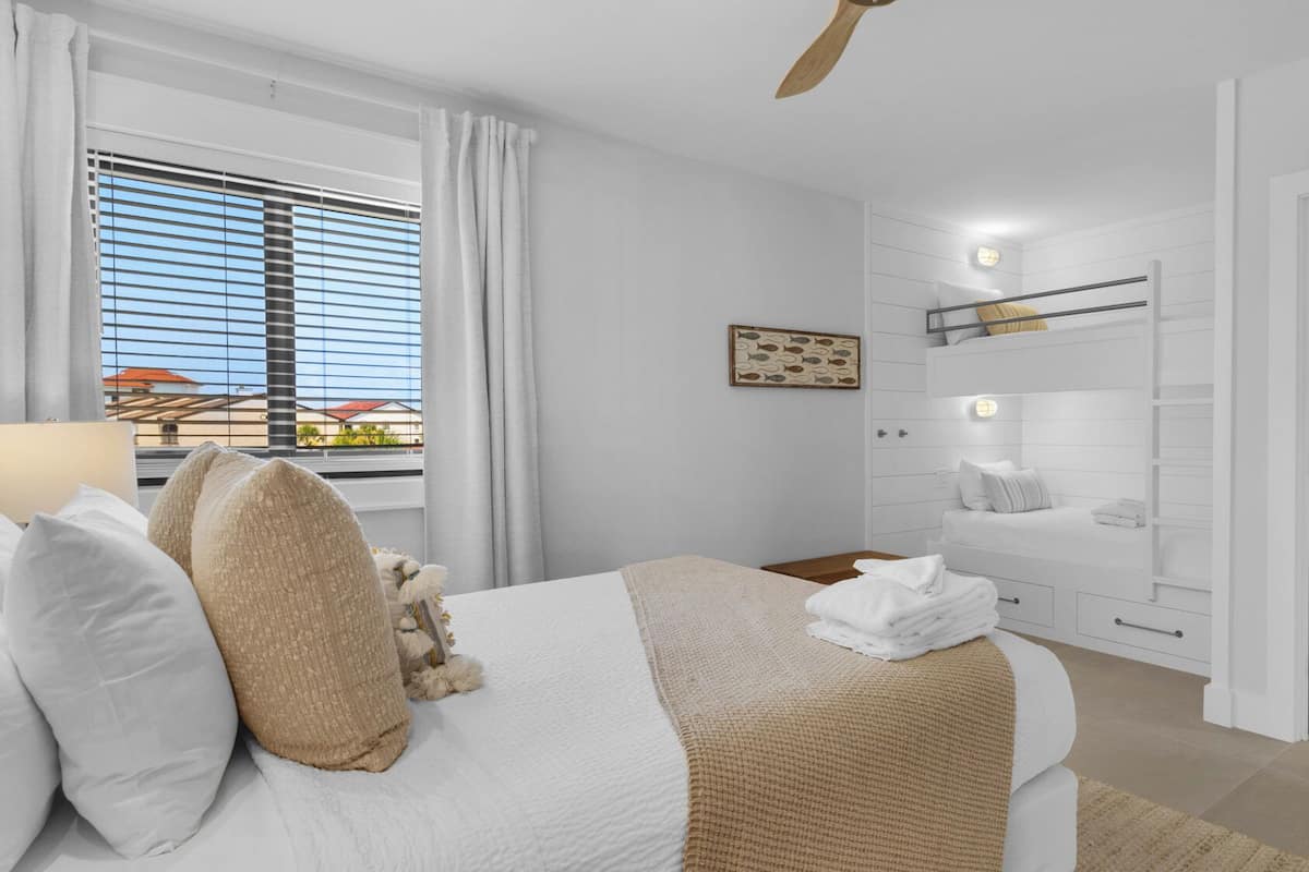 A modern bedroom with a bed, a bedside table, a nightstand, a window with blinds, a ceiling fan, a bathroom with a sink and mirror, and a view of buildings outside.