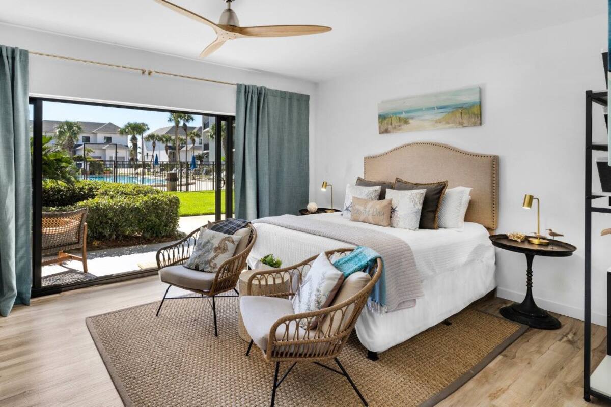 Una habitación de hotel moderna con una cama grande, un balcón con vistas a una piscina y edificios, mobiliario de mimbre y un ventilador de techo.