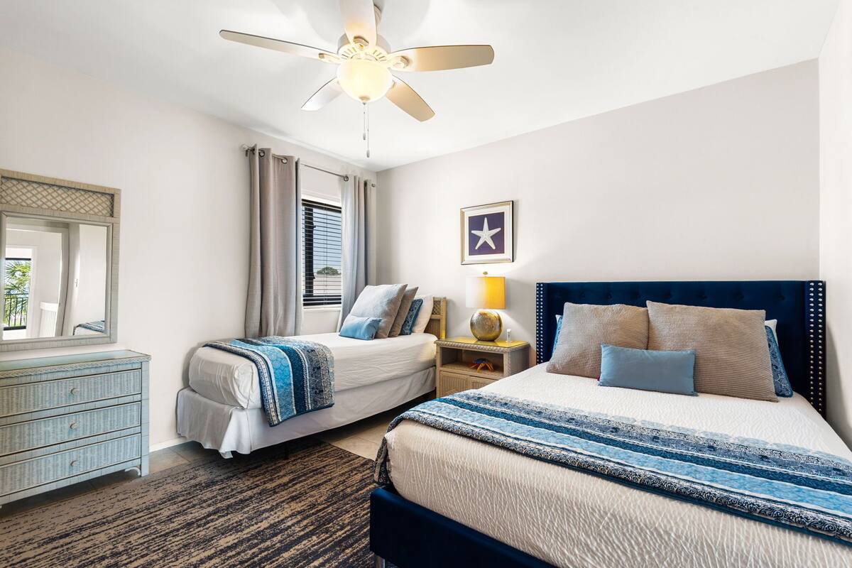 A hotel room with two beds, a ceiling fan, a nightstand with a lamp, a mirror, and a framed picture on the wall.