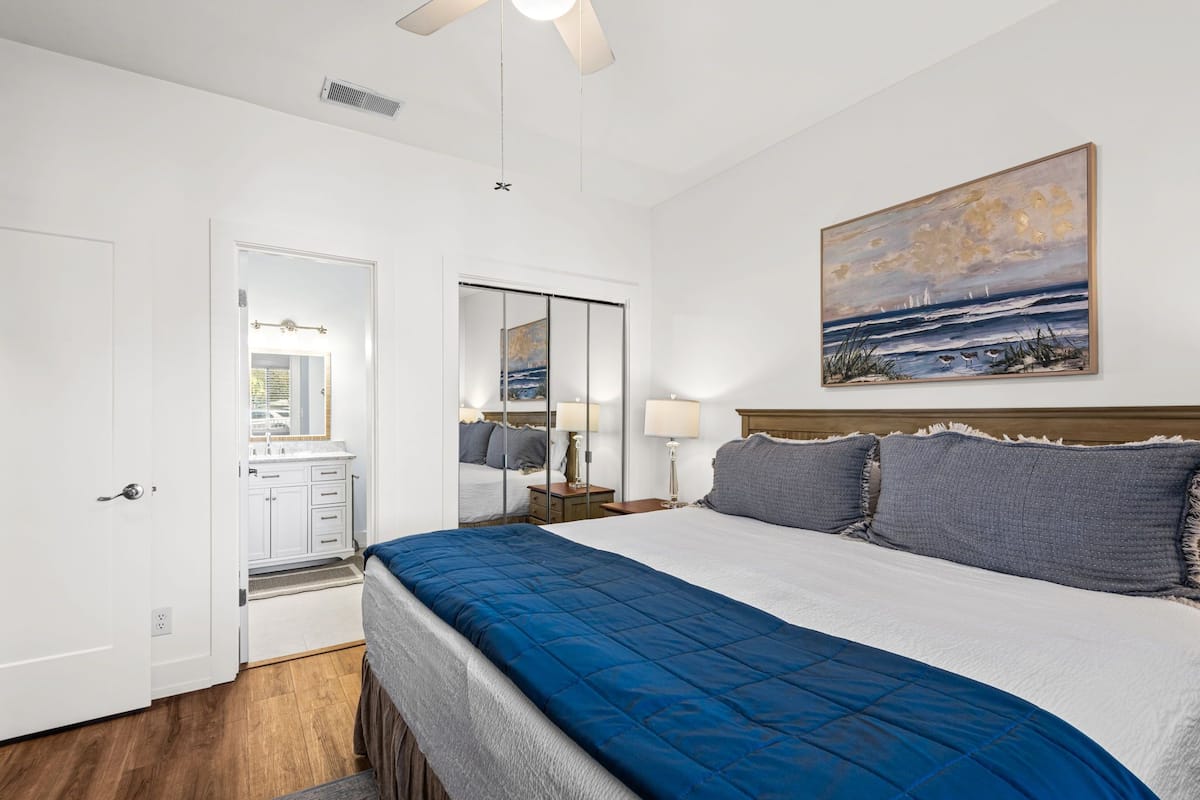A bedroom with a bed, a nightstand, a lamp, a painting, a door, and a mirror.