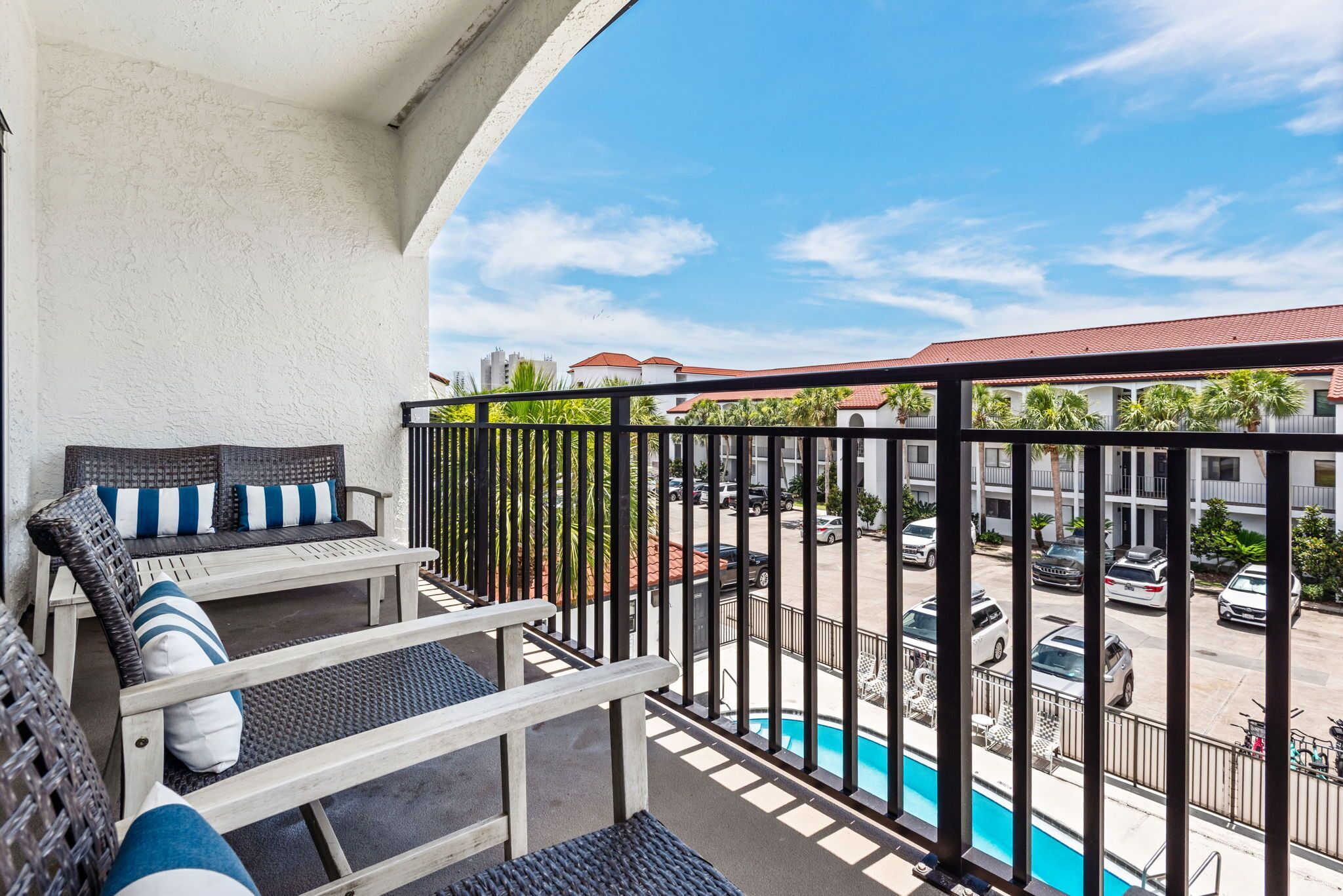 A balcony with wicker furniture, a pool, and a view of a parking lot and buildings.