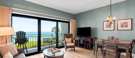 A spacious living room with a large sliding glass door leading to a balcony, a television, a dining area, and a ceiling fan.