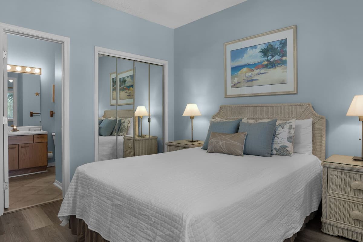 A bedroom with a large bed, two bedside lamps, a painting above the bed, and a bathroom visible through an open door.