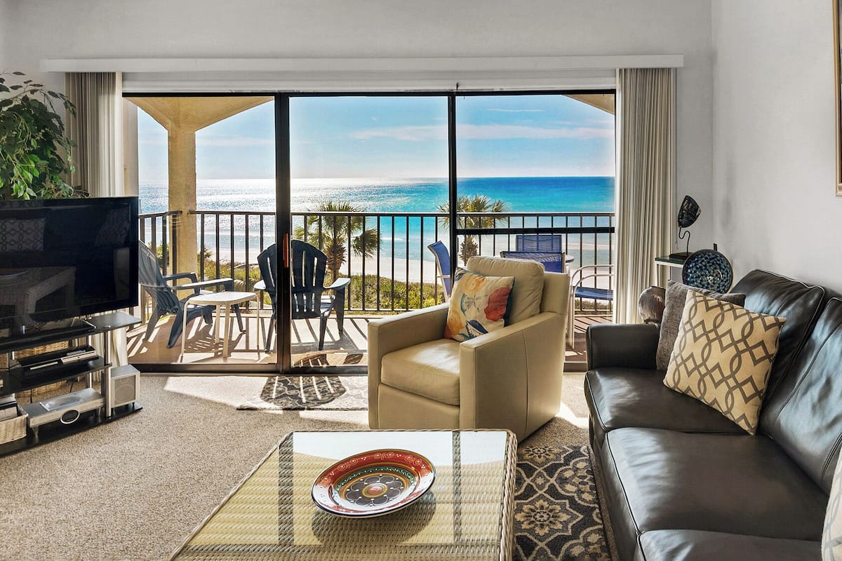 A living room with a leather sofa, a beige armchair, and a television. There is a balcony with a view of the ocean.