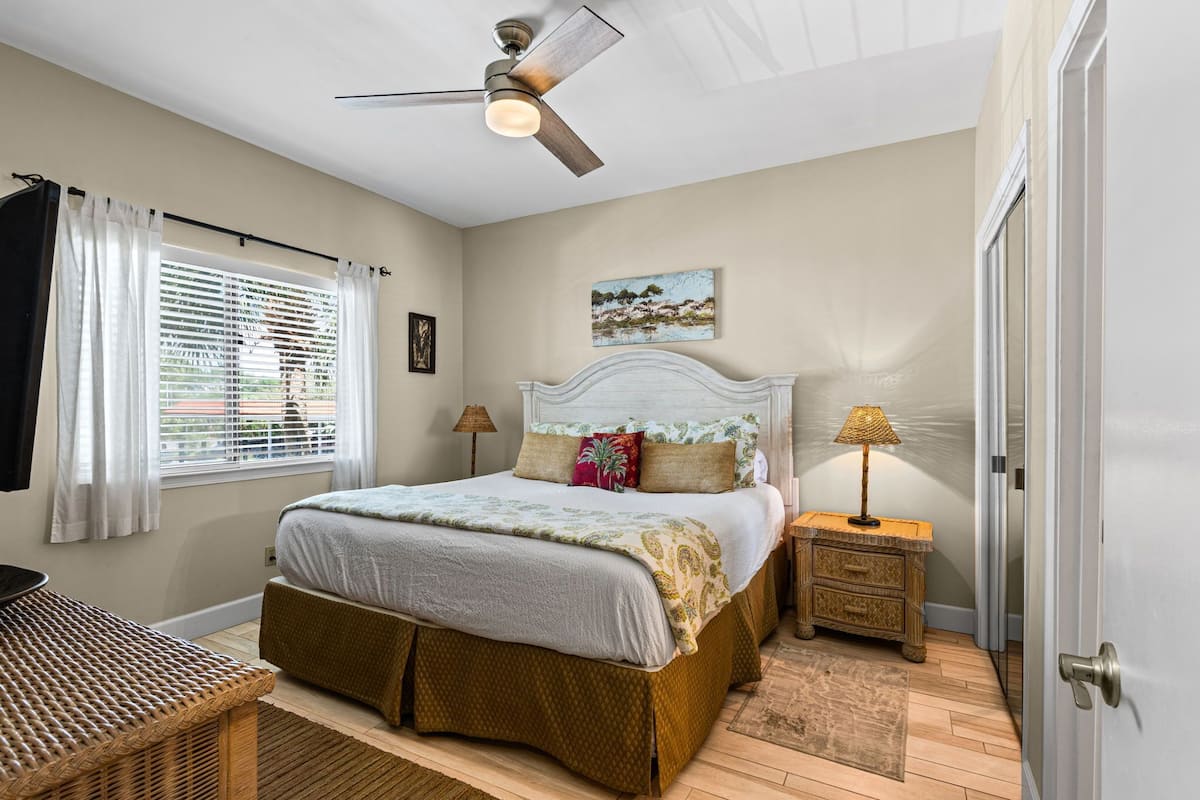 A bedroom with a bed, a ceiling fan, a nightstand with a lamp, a window with blinds, and a door.