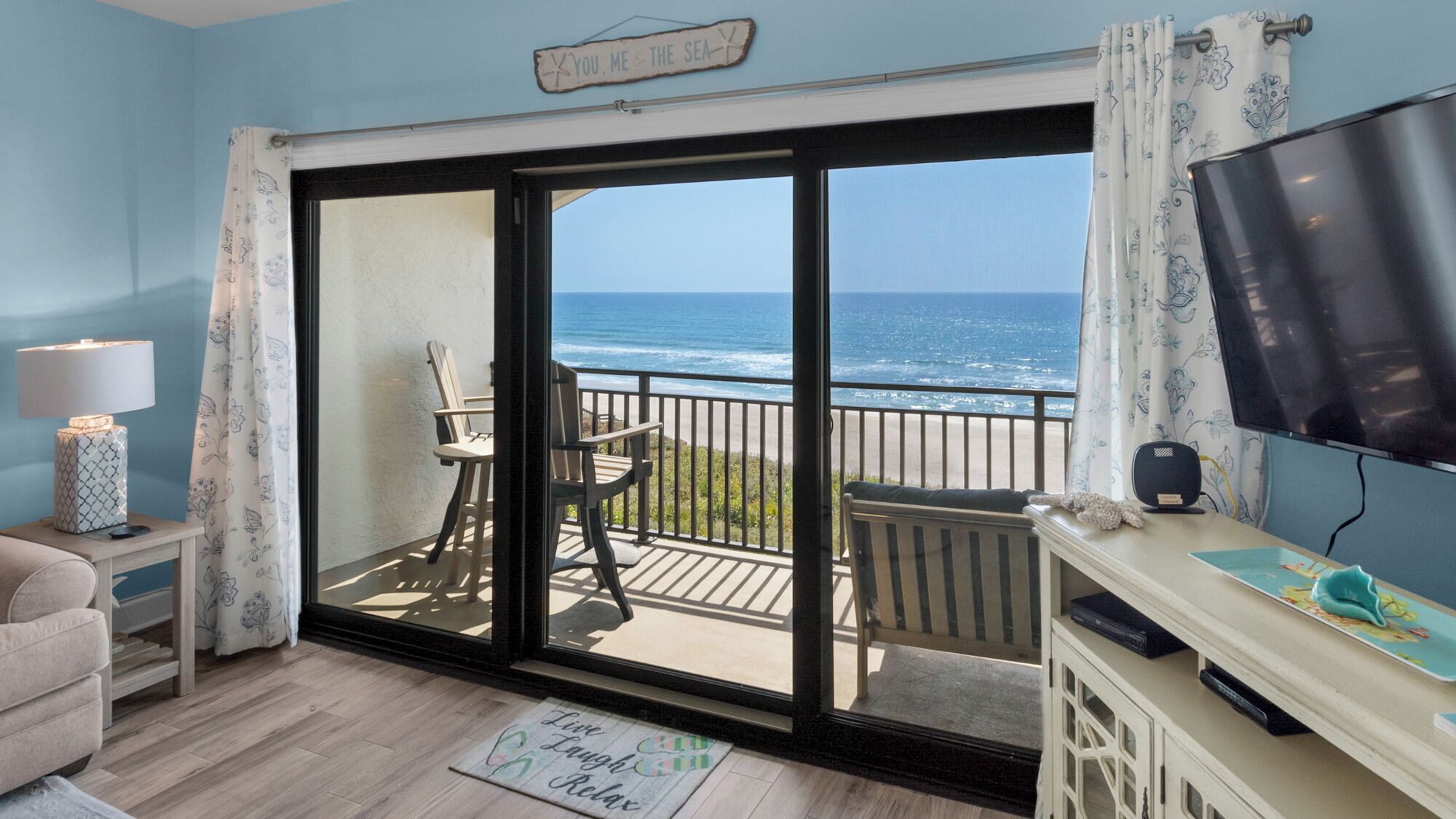 A room with a large sliding glass door leading to a balcony with a view of the ocean. There is a chair and a small table on the balcony, a lamp on a side table, and a television on a stand.