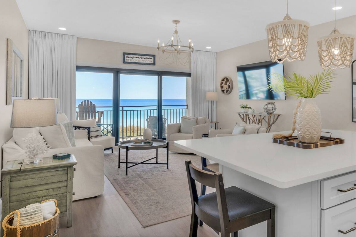 A spacious living area with a large window overlooking the ocean, a central island with a vase, and a modern kitchen with white cabinetry.