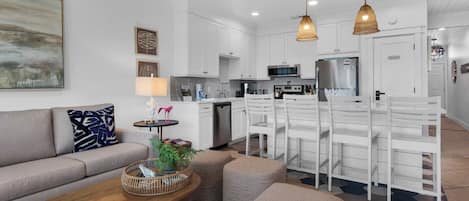 A modern kitchen with white cabinets, a stainless steel refrigerator, and a microwave. There is a dining area with white chairs and a table.
