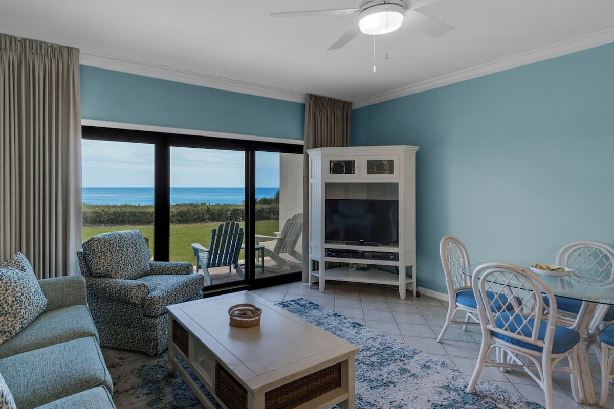 A living room with a sofa, chairs, a coffee table, a TV stand, and a view of the ocean.