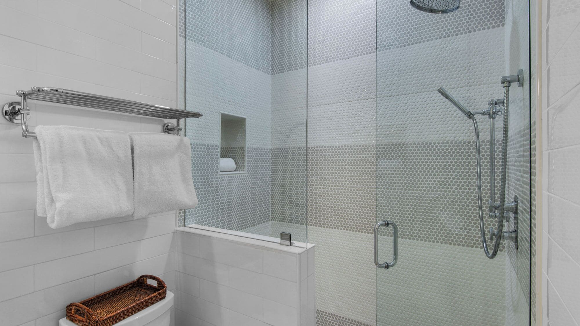 Une salle de bain moderne avec une douche à parois de verre, des murs recouverts de carreaux blancs, un porte-serviettes et un panier en osier.