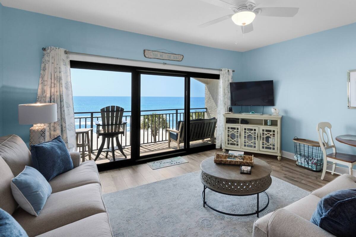 A living room with a view of the sea, a ceiling fan, a TV, and a dining area with a table and chairs.