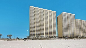 Ein Hotel direkt am Strand mit mehreren Balkonen, ein sandiger Strand im Vordergrund und ein klarer, blauer Himmel.