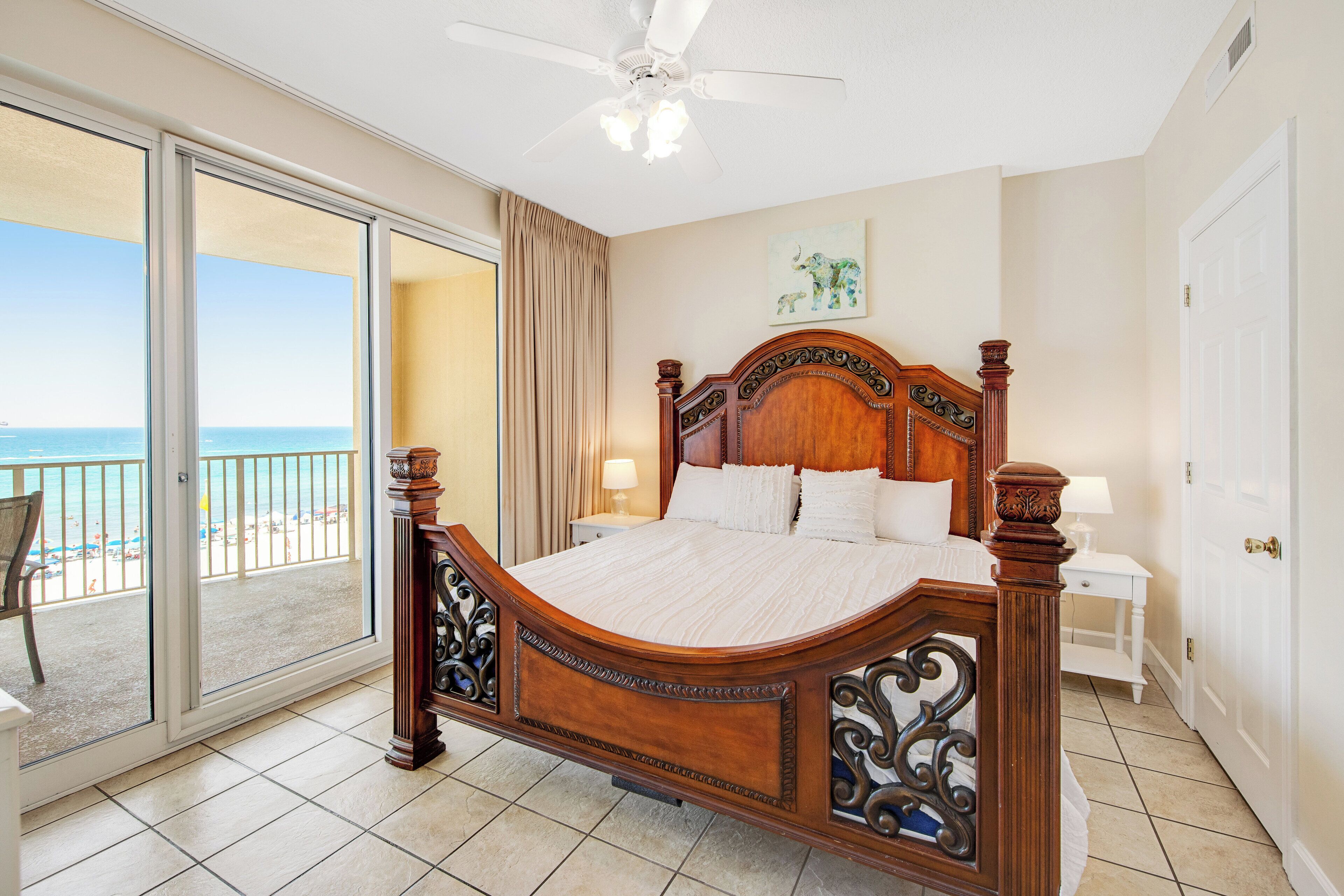 Un dormitorio con una cama grande, un ventilador de techo, vista a la playa y puerta corrediza al balcón.