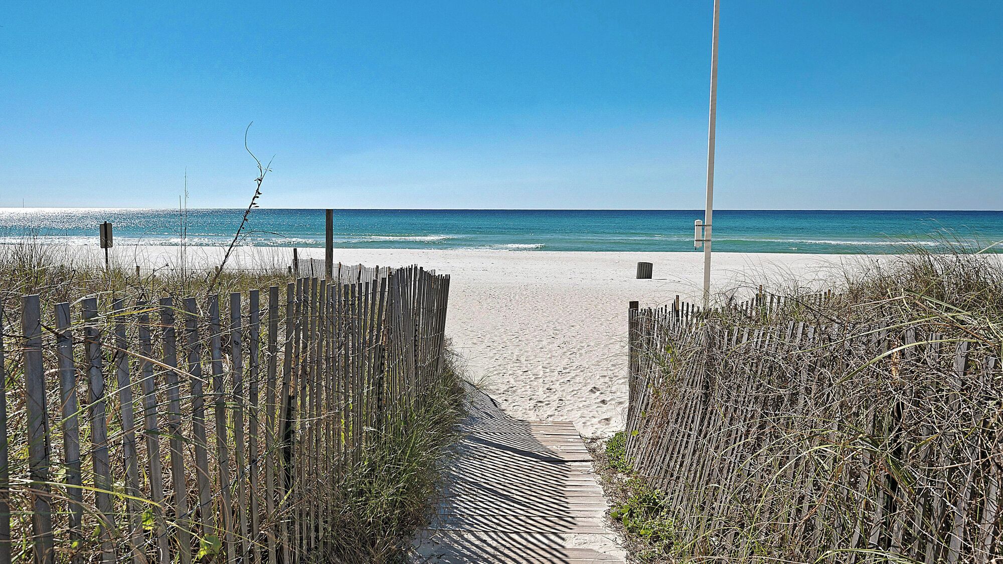 A sandy beach path leading to the ocean, flanked by wooden fences and a clear blue sky.