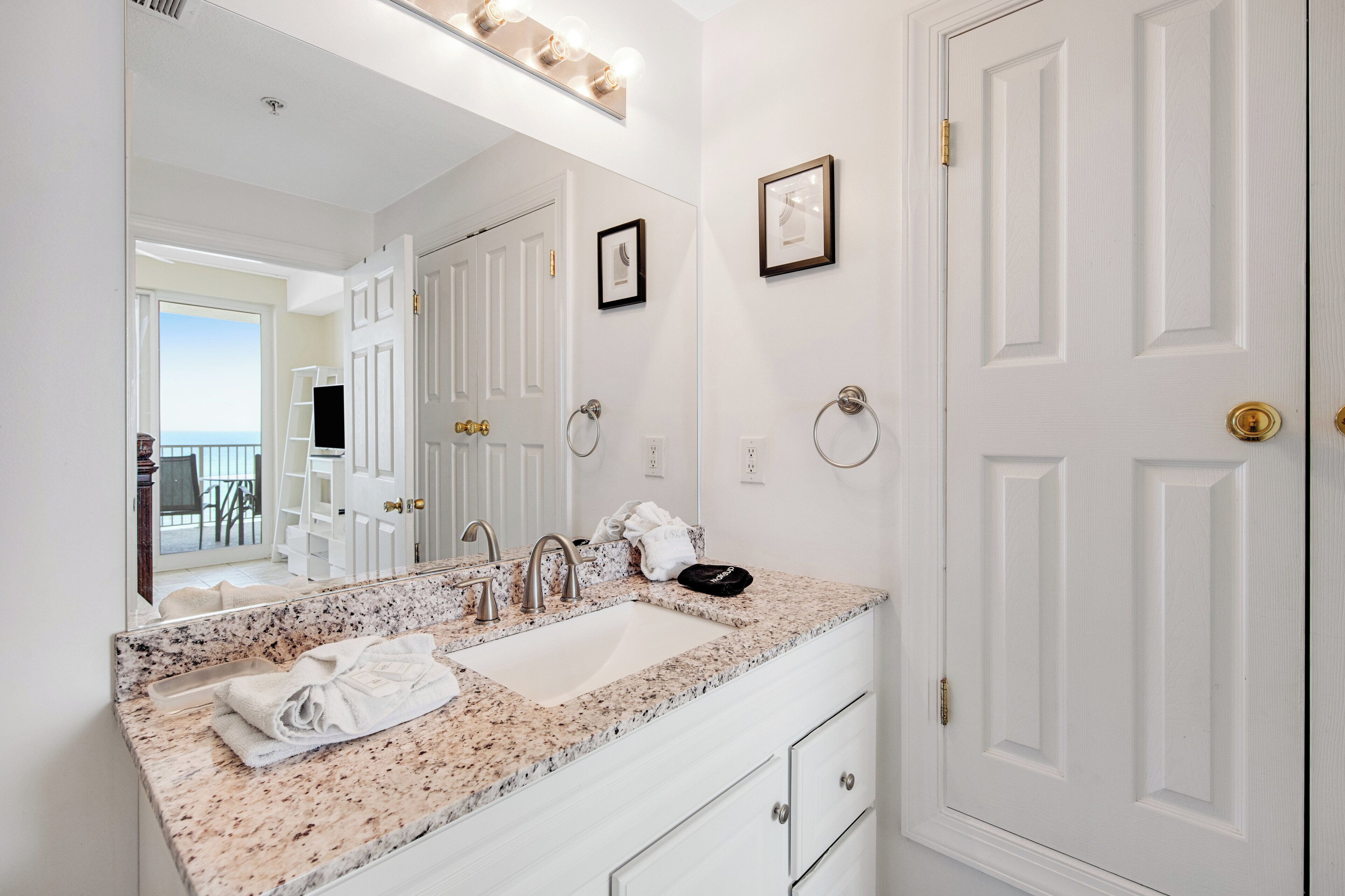 A bathroom with a large mirror, a sink, and a vanity. There are two framed pictures on the wall and a door with a brass handle.