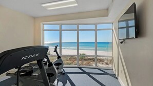 Ein Zimmer mit großem Fenster und Blick auf den Strand, einem Laufband und einem an der Wand befestigten Flachbildfernseher.
