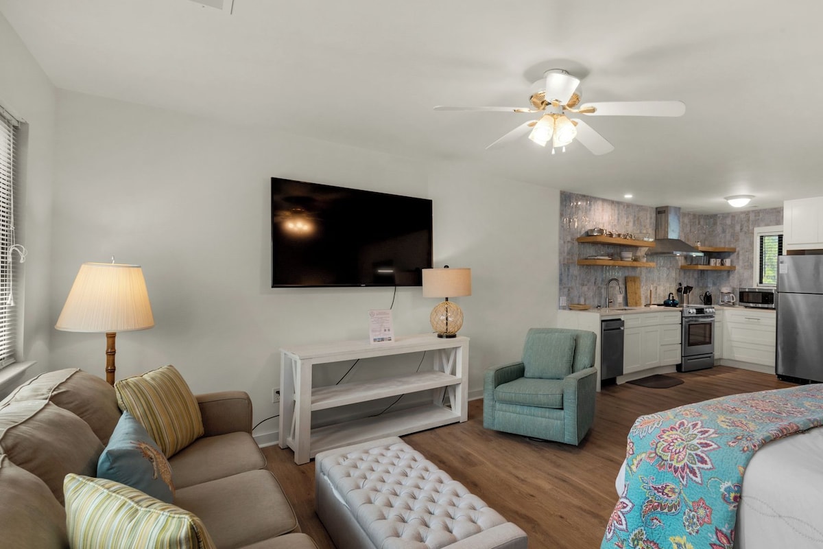 A compact living space with a sofa, armchair, and a bed with a patterned bedspread. A television is mounted on the wall, and a ceiling fan is visible.
