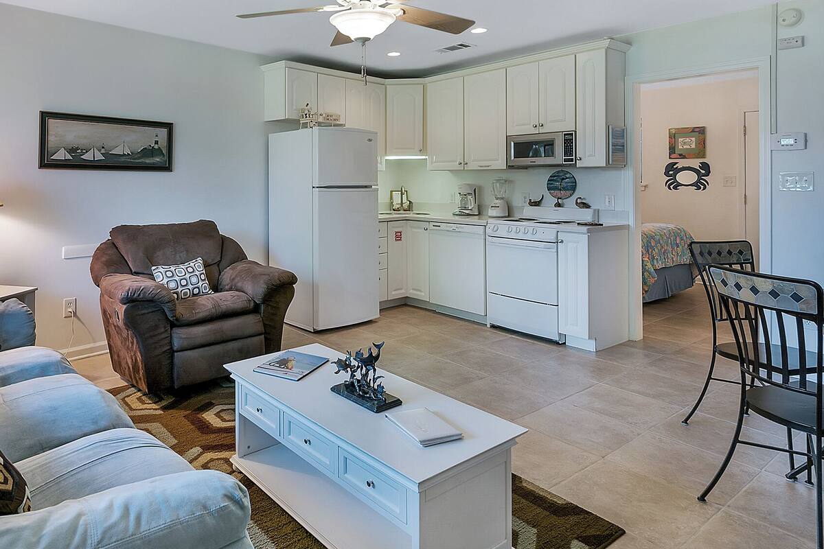 A modern living room with a sofa, armchair, and coffee table. A dining area with a table and chairs is adjacent to the kitchen, which includes white cabinets, a microwave, and a stove.