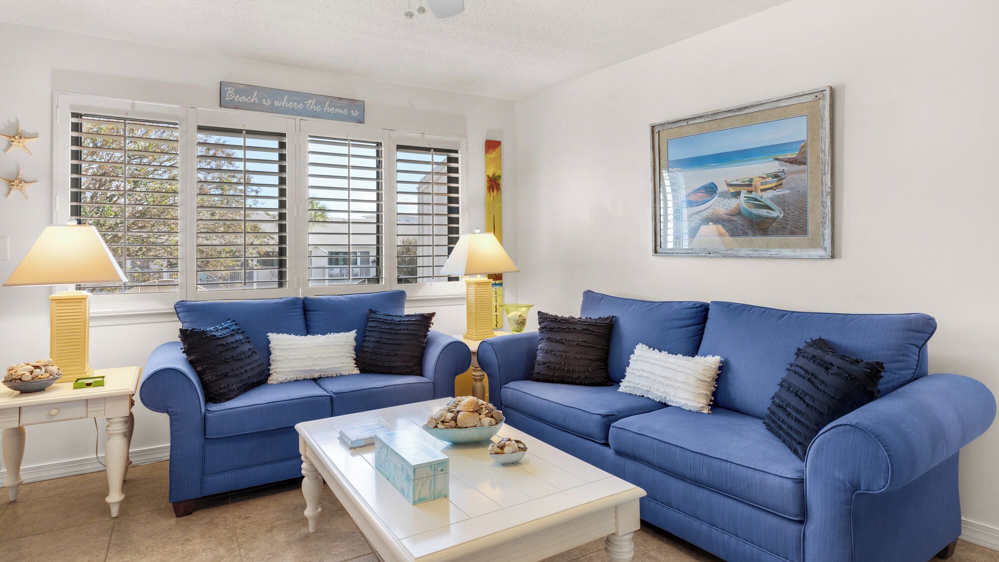 Ein Wohnzimmer mit zwei blauen Sofas, einem weißen Couchtisch und einem Bild mit einer Strandansicht.