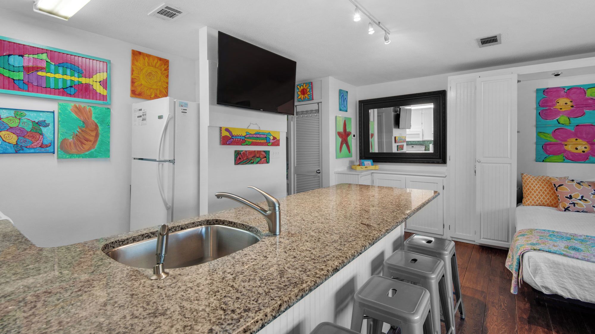 A modern kitchen with granite countertops, stainless steel sink, and a built-in oven. There are colorful artwork on the walls and a large mirror above the sink.