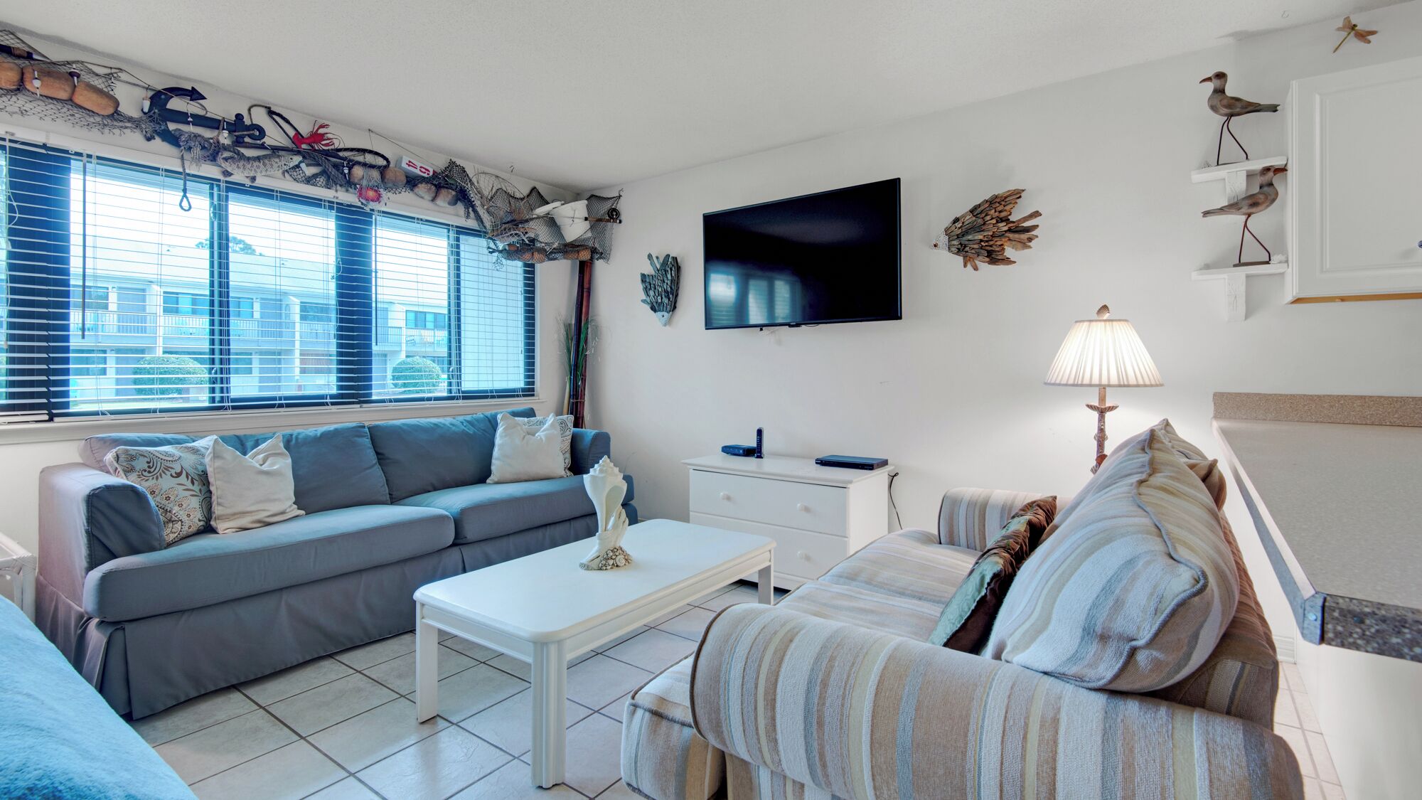 A modern living room with a grey sofa, a white coffee table, and a striped armchair. A flat-screen TV is mounted on the wall, and there are decorative wall hangings.