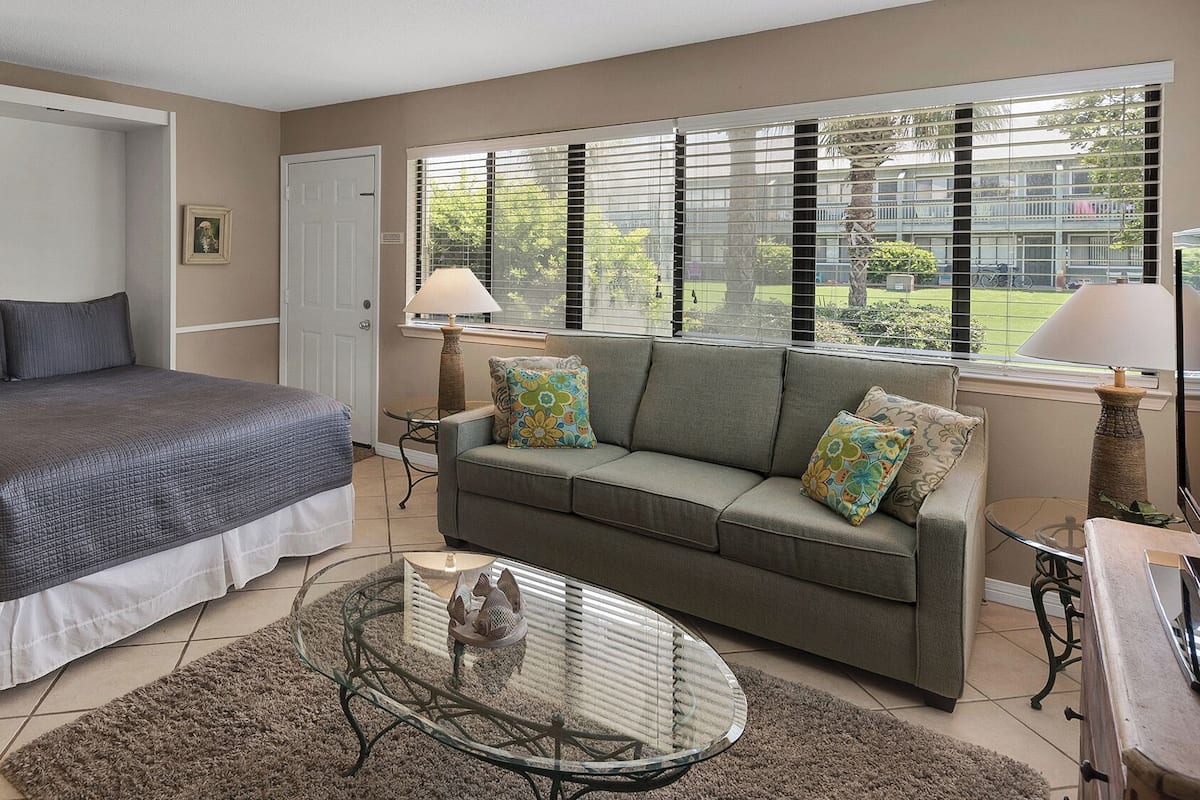A bedroom with a bed, a sofa, a glass coffee table, and a window with blinds.