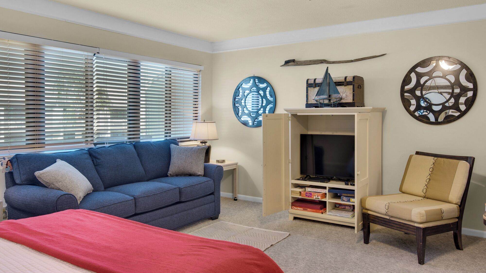 Een woonkamer met een blauwe bank, een rode plaid, een beige fauteuil, een tv-meubel met een flatscreen-tv, en decoratieve wandkunst.