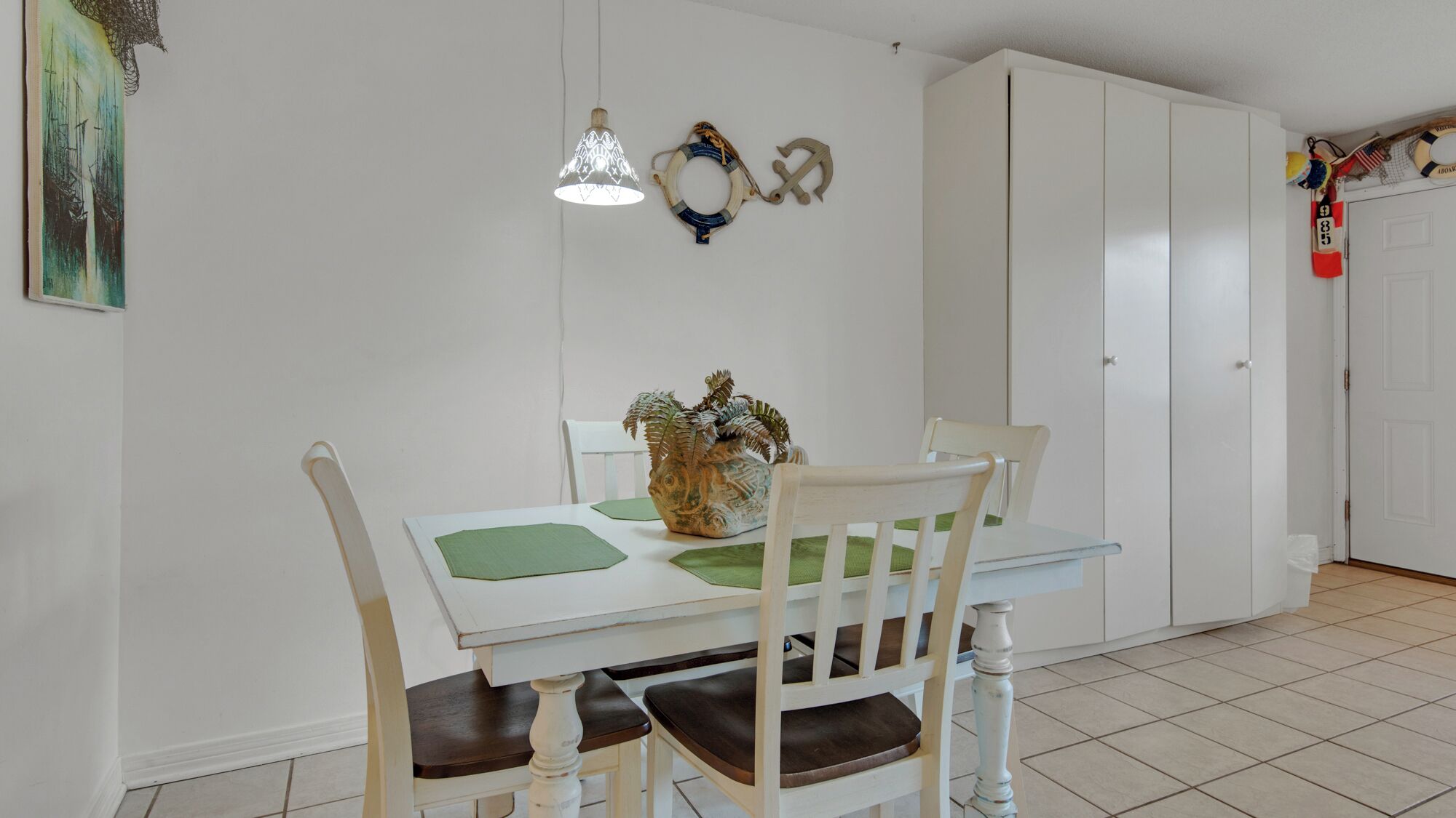 A dining area with a white table, green placemats, and a decorative vase. There are two hanging lamps and a white wardrobe in the background.