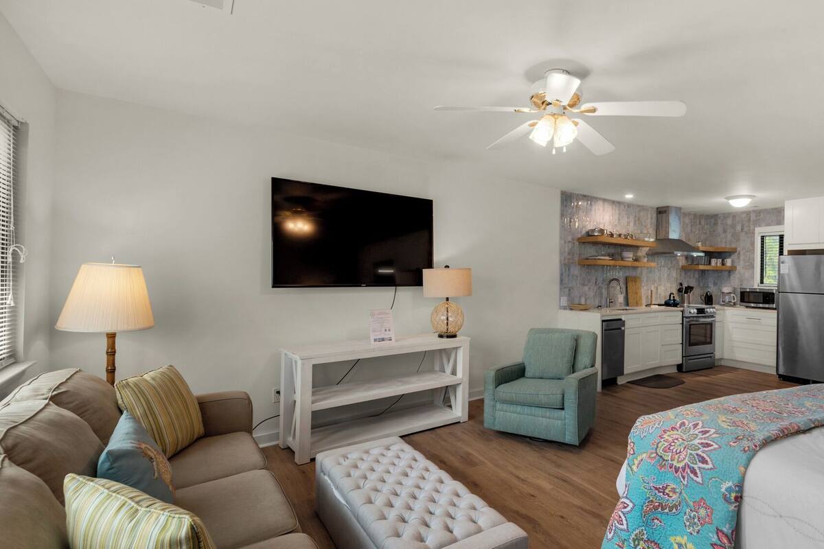 A compact living space with a sofa, armchair, and a bed with a patterned bedspread. A television is mounted on the wall, and a ceiling fan is visible.