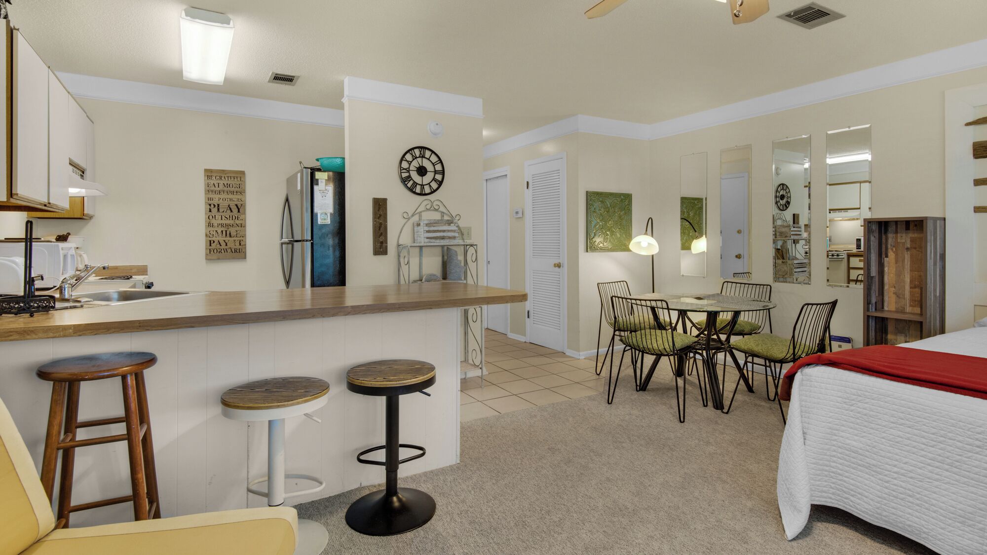Una habitación de hotel compacta con kitchenette, un comedor con mesa y sillas, una cama con una colcha roja, y un baño con ducha e inodoro.