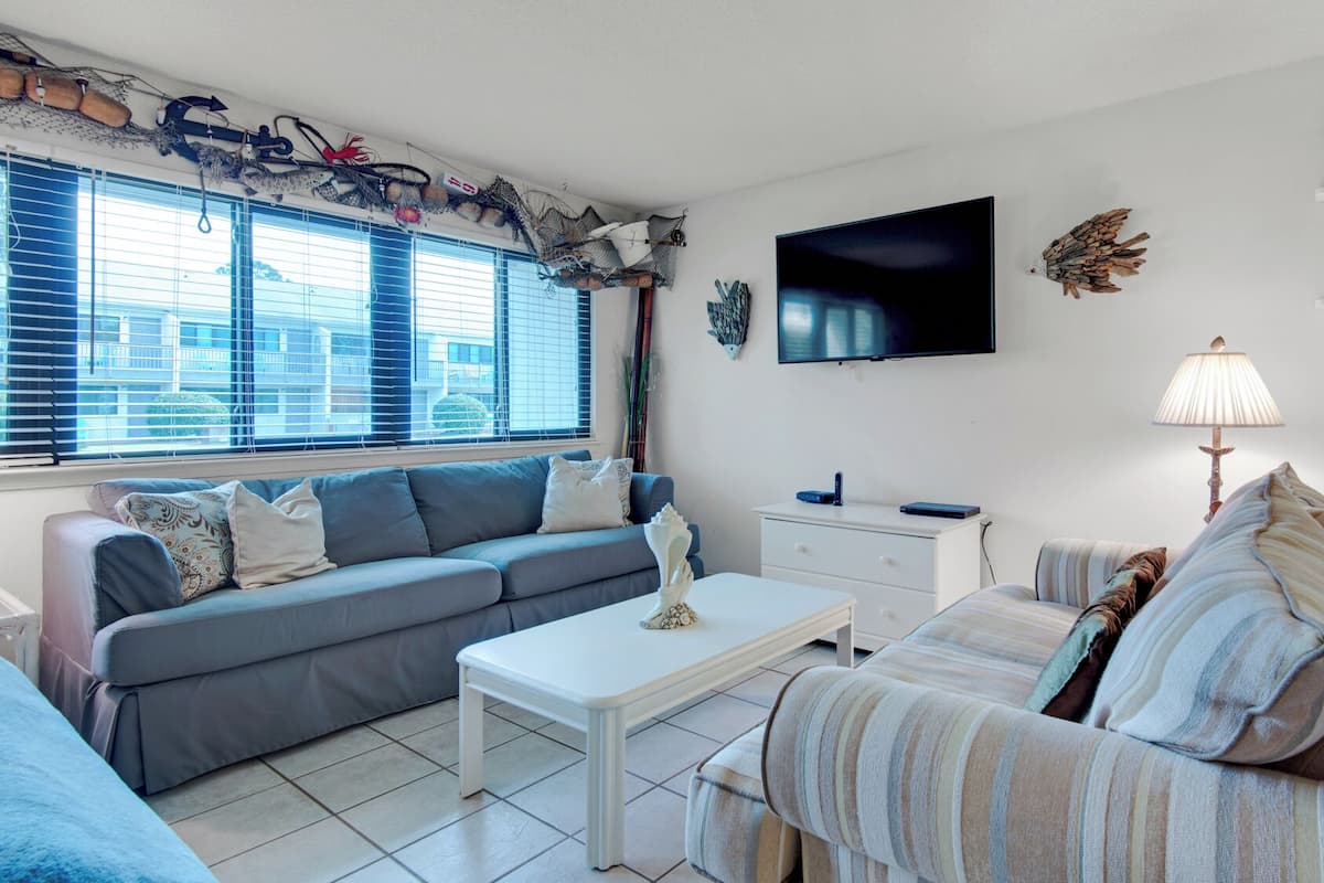 A modern living room with a grey sofa, a white coffee table, and a striped armchair. A flat-screen TV is mounted on the wall, and there are decorative wall hangings.