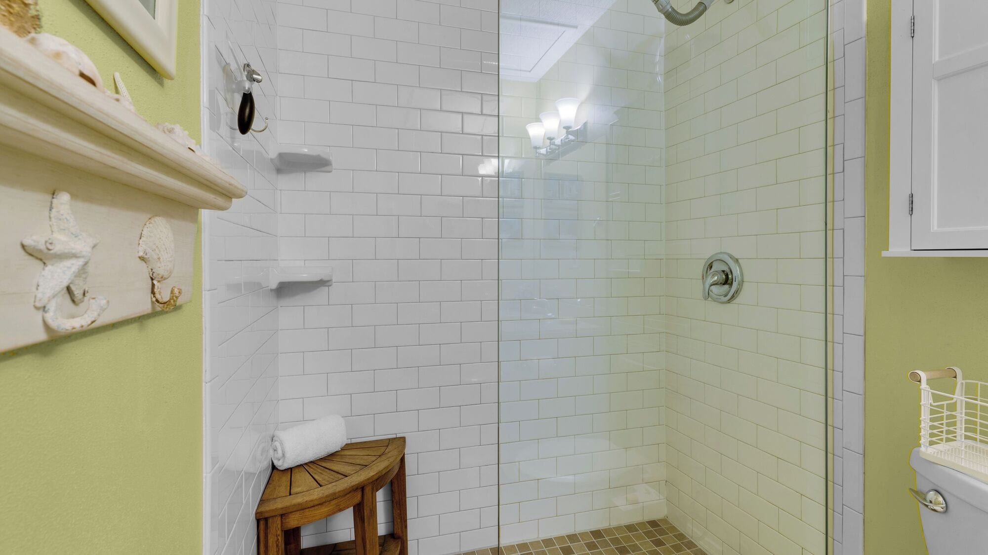Une salle de bain avec des carreaux de céramique blancs rectangulaires, un tabouret en bois, une serviette et une cabine de douche en verre.