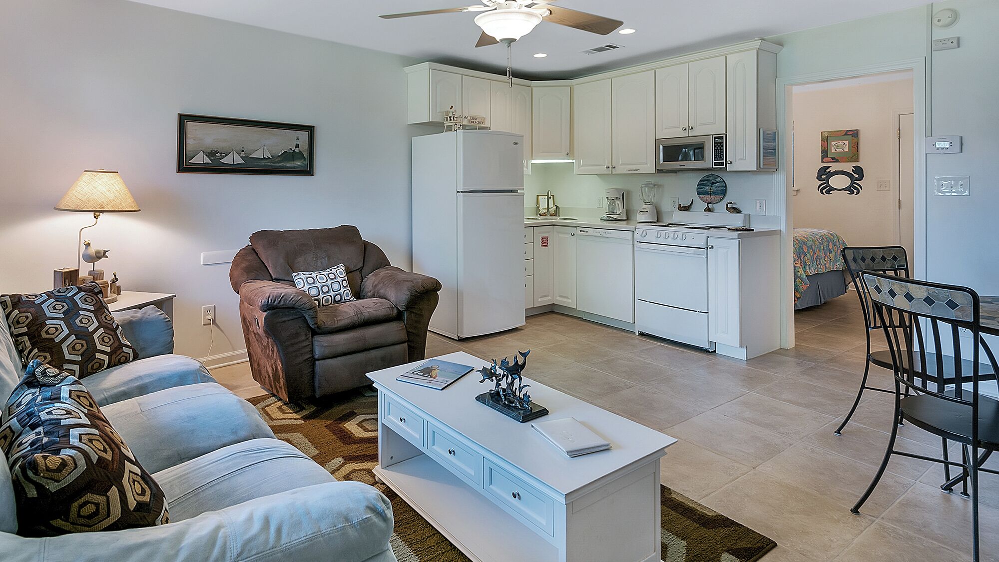 A modern living room with a sofa, armchair, and coffee table. A dining area with a table and chairs is adjacent to the kitchen, which includes white cabinets, a microwave, and a stove.