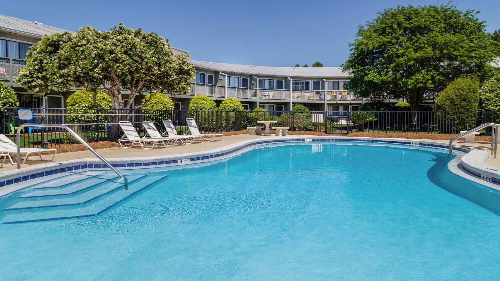 Une piscine dotée de chaises longues et entourée d’une clôture, le tout bordé d’arbres et adjacent à un bâtiment avec des balcons.