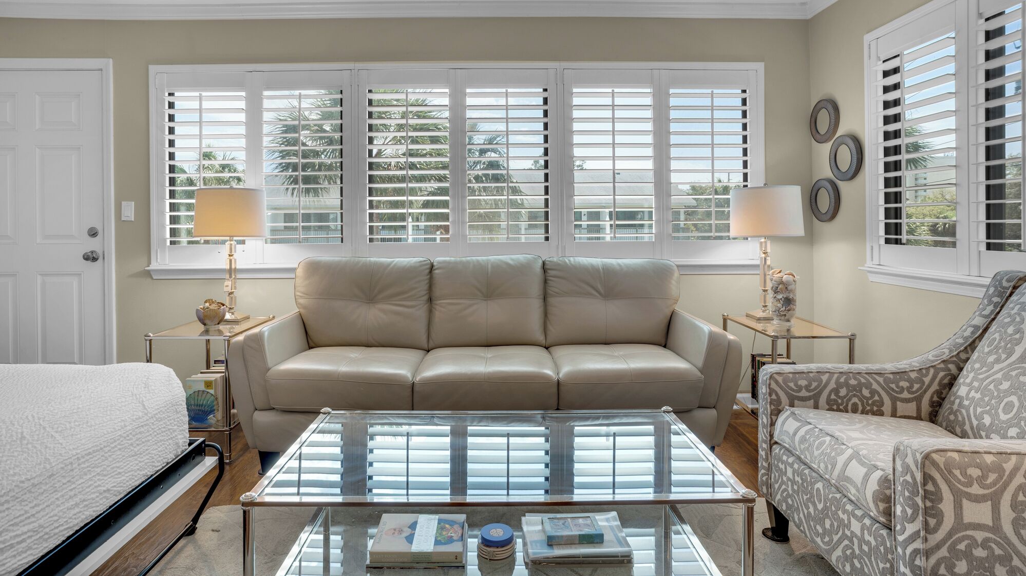 A living room with a beige sofa, a patterned armchair, a glass coffee table, and a view through large windows with shutters.