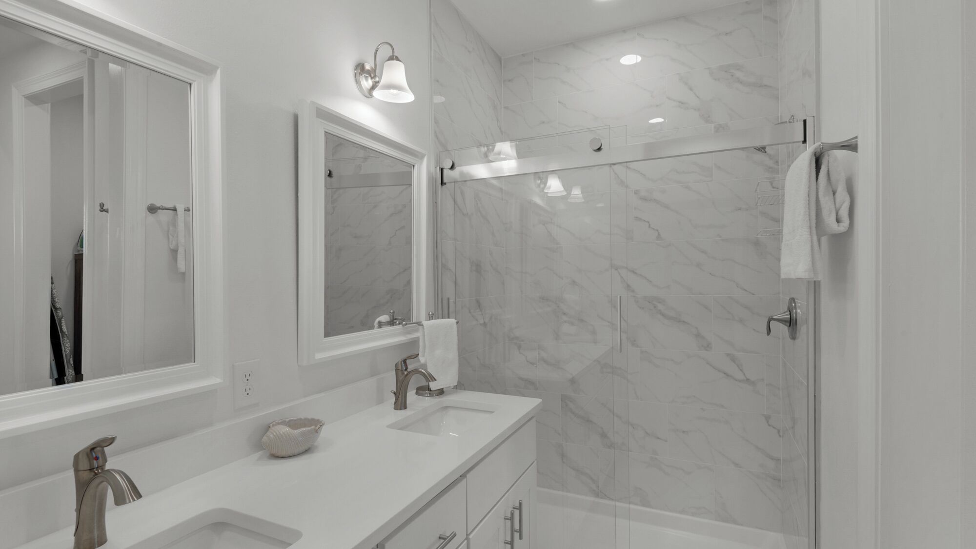 A modern bathroom with a large mirror, two sinks, a shower, and marble walls.