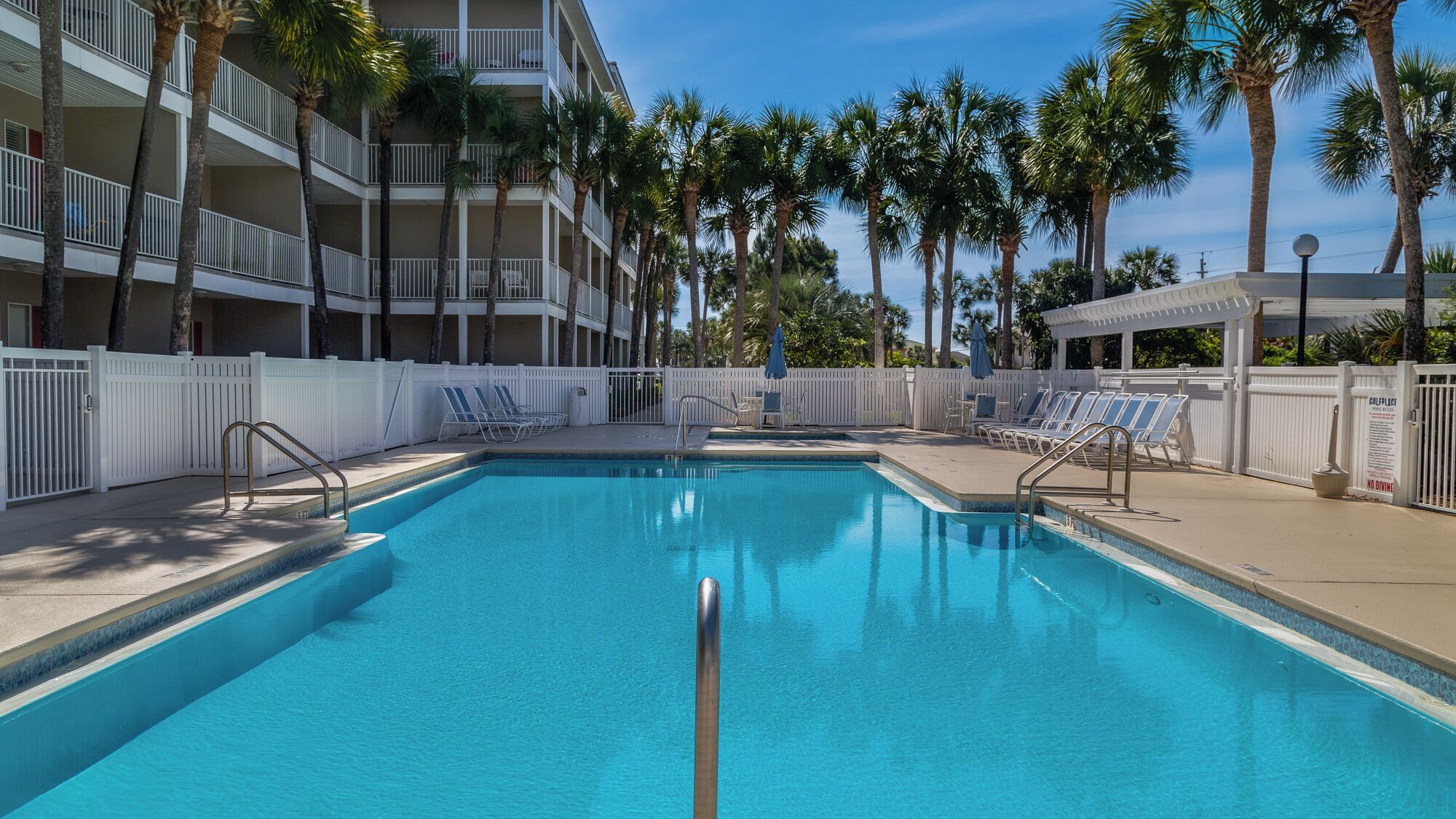 A swimming pool with lounge chairs, surrounded by palm trees and a multi-story building with balconies.