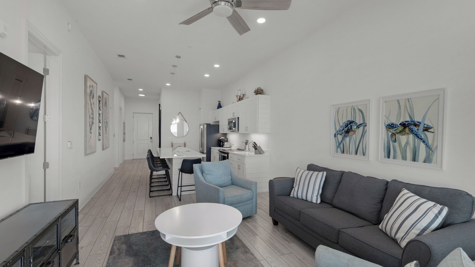 Una sala de estar moderna con un sofá gris, una mesa de centro blanca y un sillón azul. Se observa una zona de cocina con armarios blancos y una mesa de comedor con sillas.