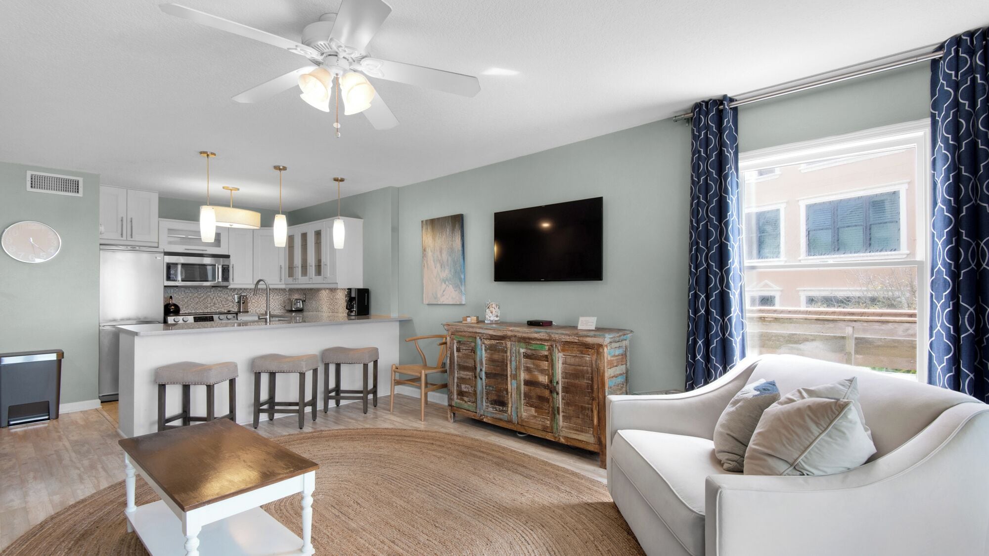 A modern living room with a white sofa, a wooden coffee table, and a kitchen area with white cabinets and a flat-screen TV.