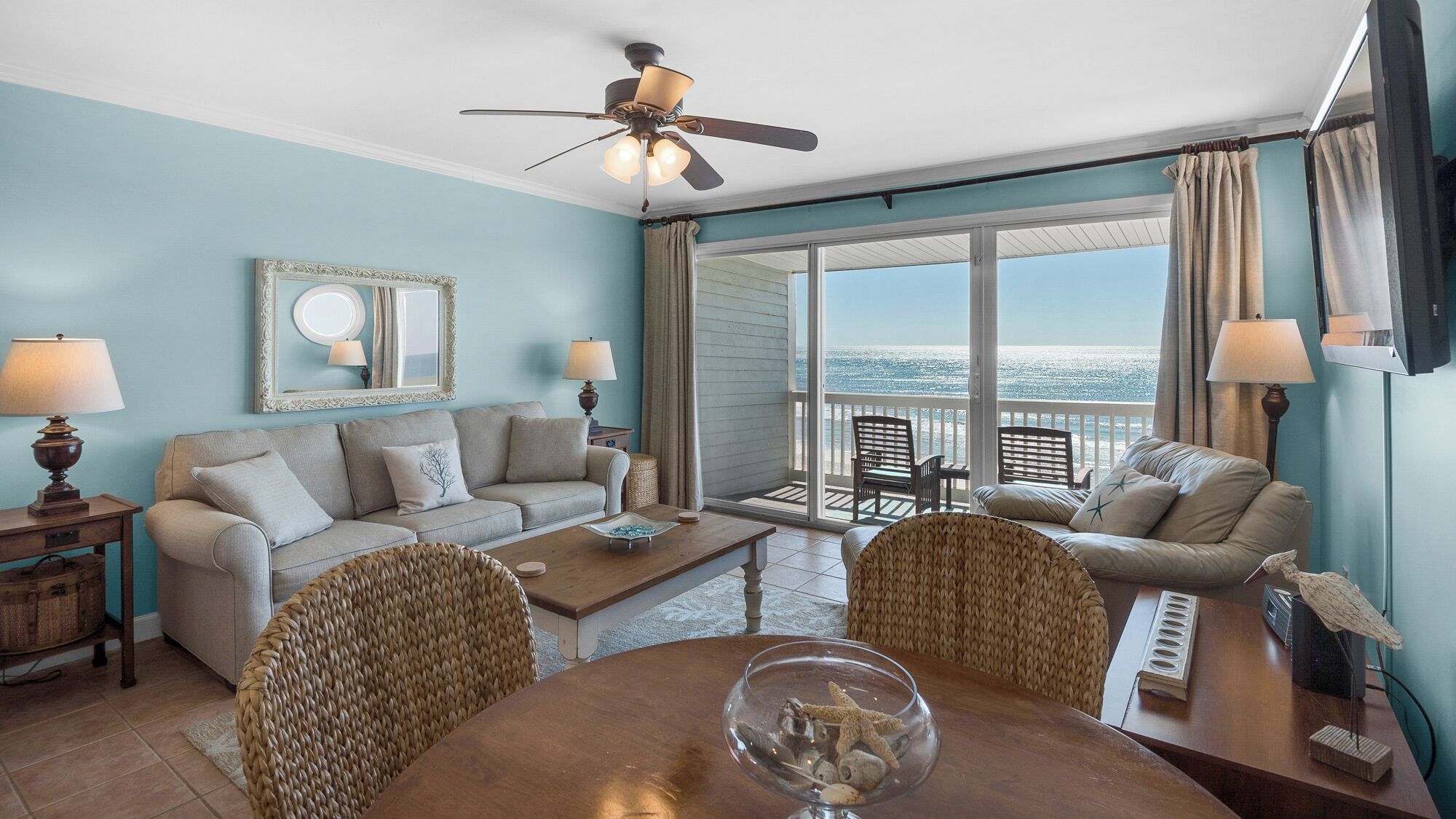 A living room with a sofa, two armchairs, a coffee table, and a TV. There is a balcony with a view of the ocean.