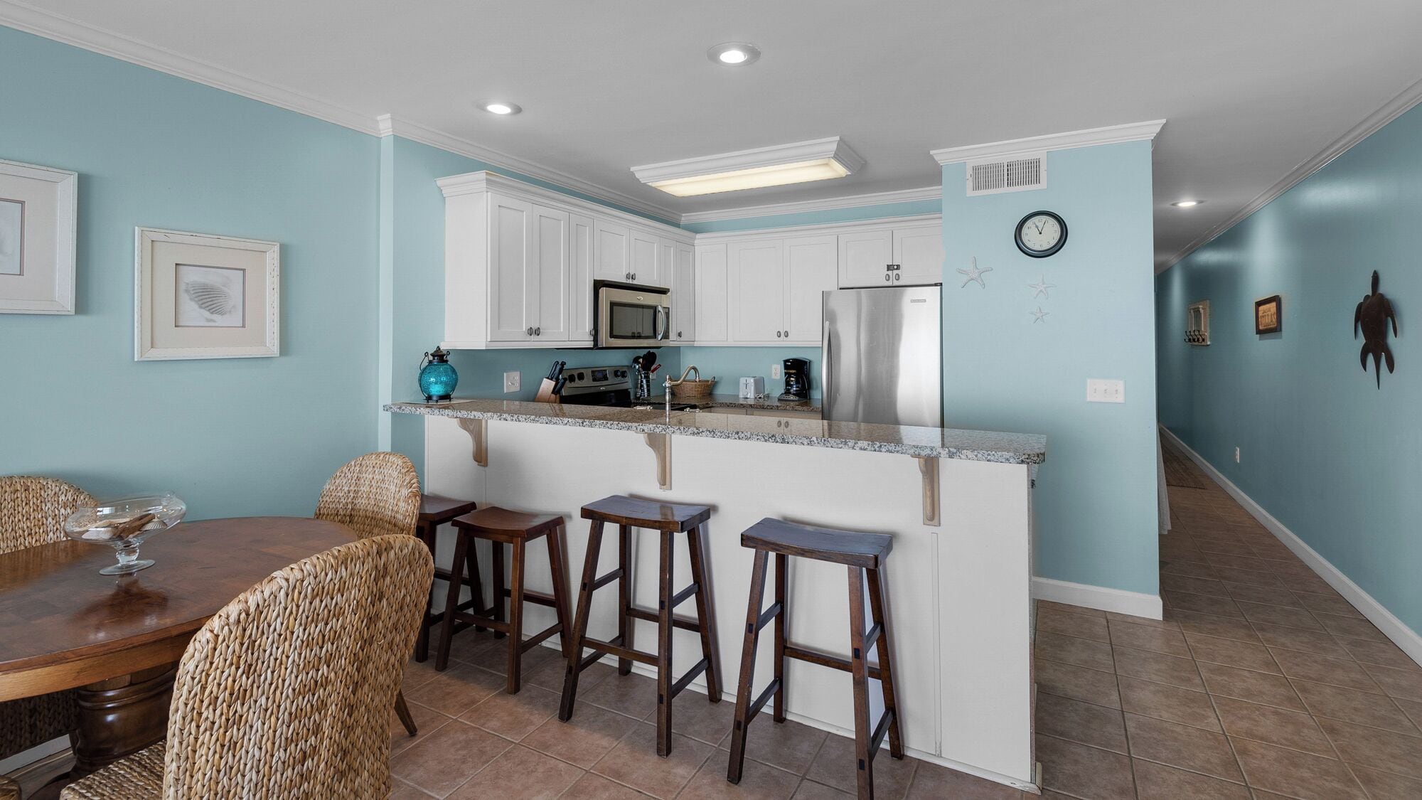 Una cocina con armarios blancos, un microondas y un refrigerador. Hay un comedor con una mesa y sillas.