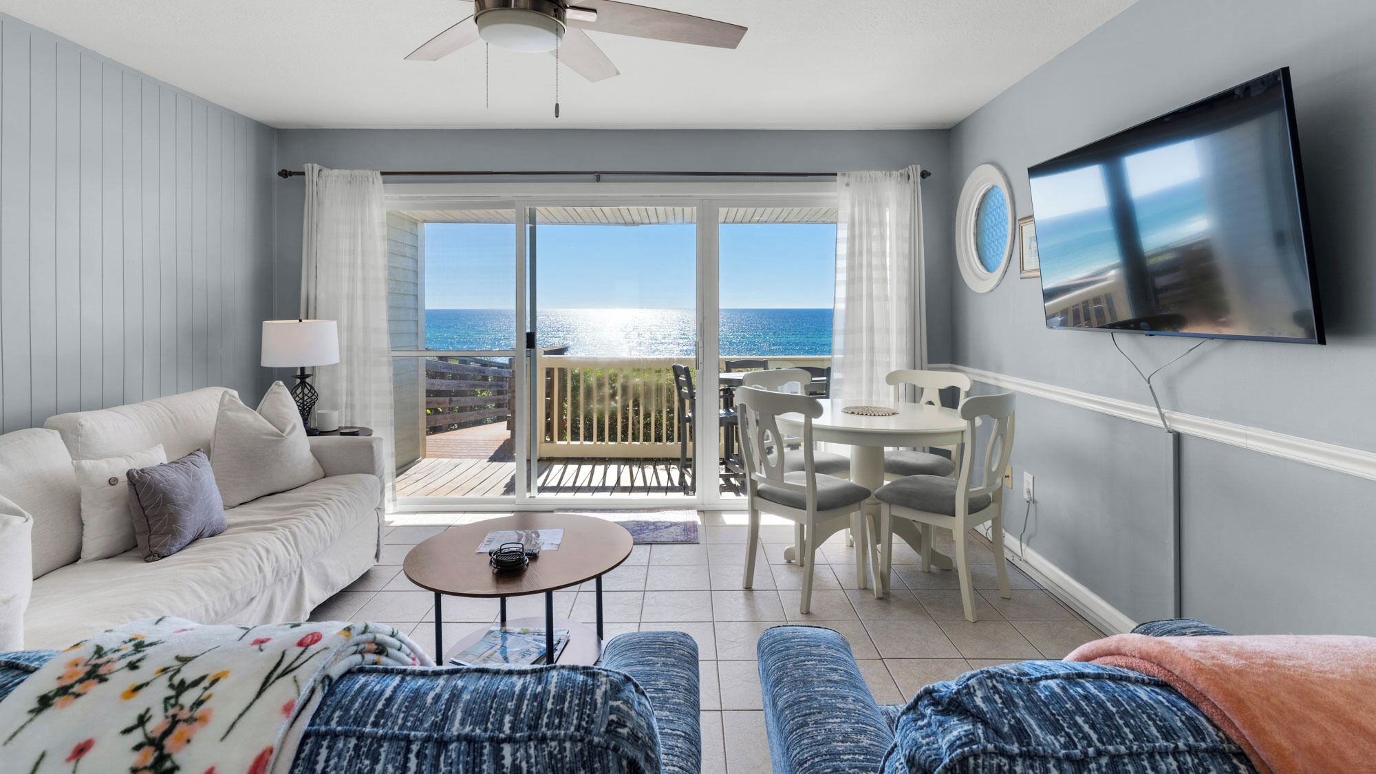 A living room with a sofa, coffee table, and ottoman, a dining area with chairs and a table, a TV mounted on the wall, and a balcony with a view of the sea.