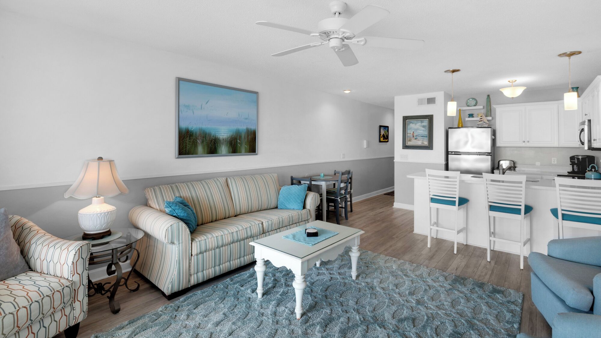 A living room with a sofa, a coffee table, and a dining area with chairs. There is a kitchen with a refrigerator and a microwave.