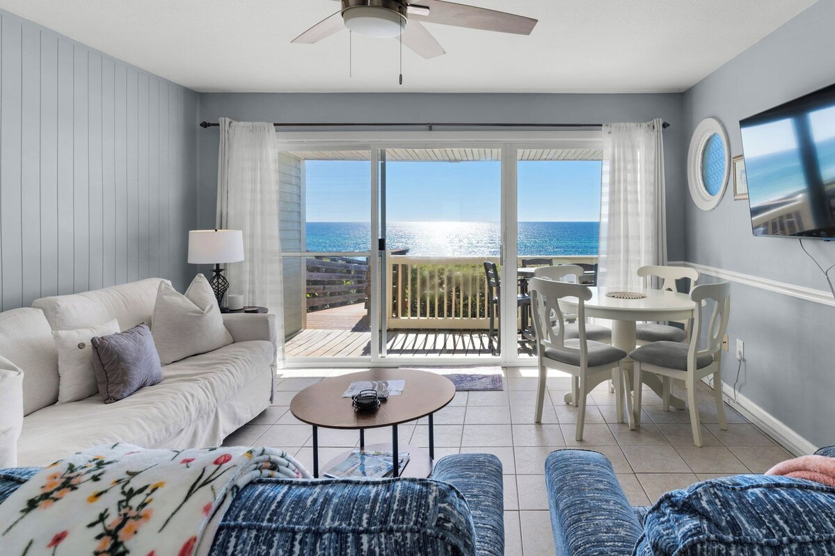 A living room with a sofa, coffee table, and ottoman, a dining area with chairs and a table, a TV mounted on the wall, and a balcony with a view of the sea.