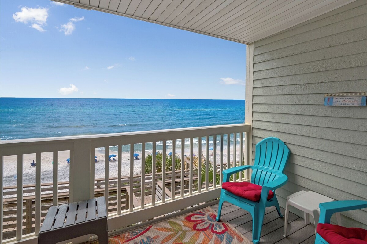 A balcony with a view of the ocean, featuring two turquoise chairs and a small table.