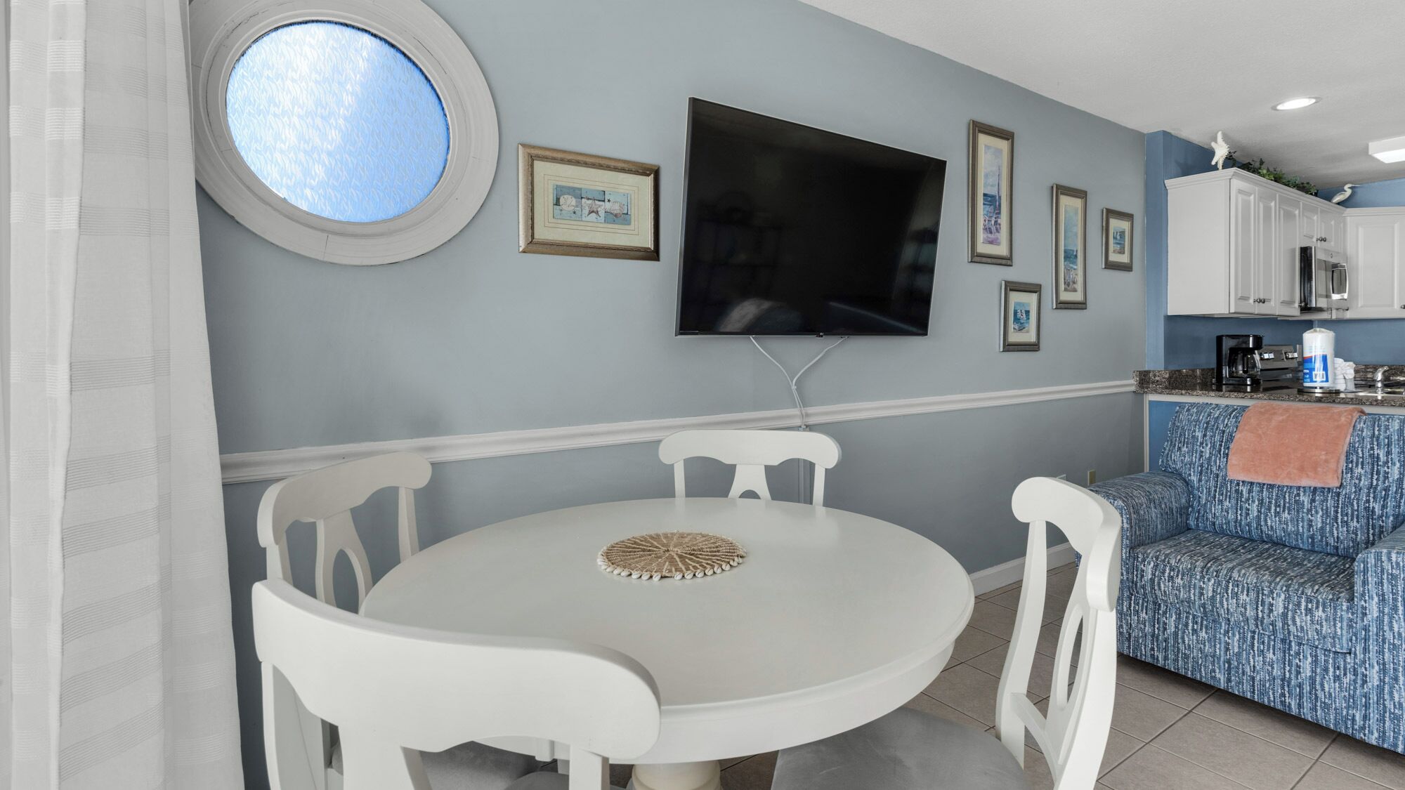 Un comedor con una mesa redonda y sillas blancas, un televisor montado en la pared y una zona de cocina con armarios y un sofá azul con estampado.