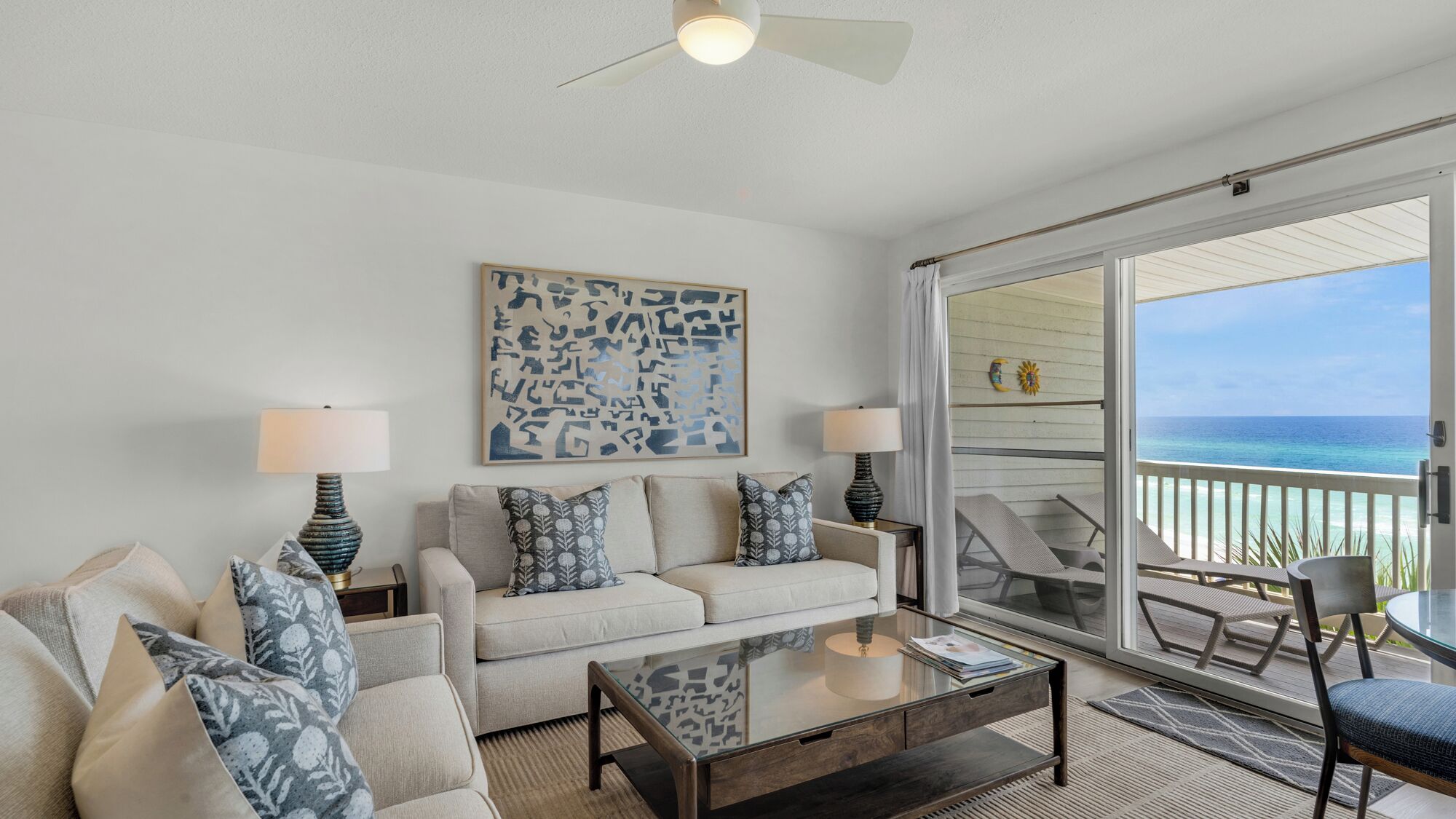 A living room with a sofa, two lamps, a coffee table, and a view of the ocean through a sliding glass door.