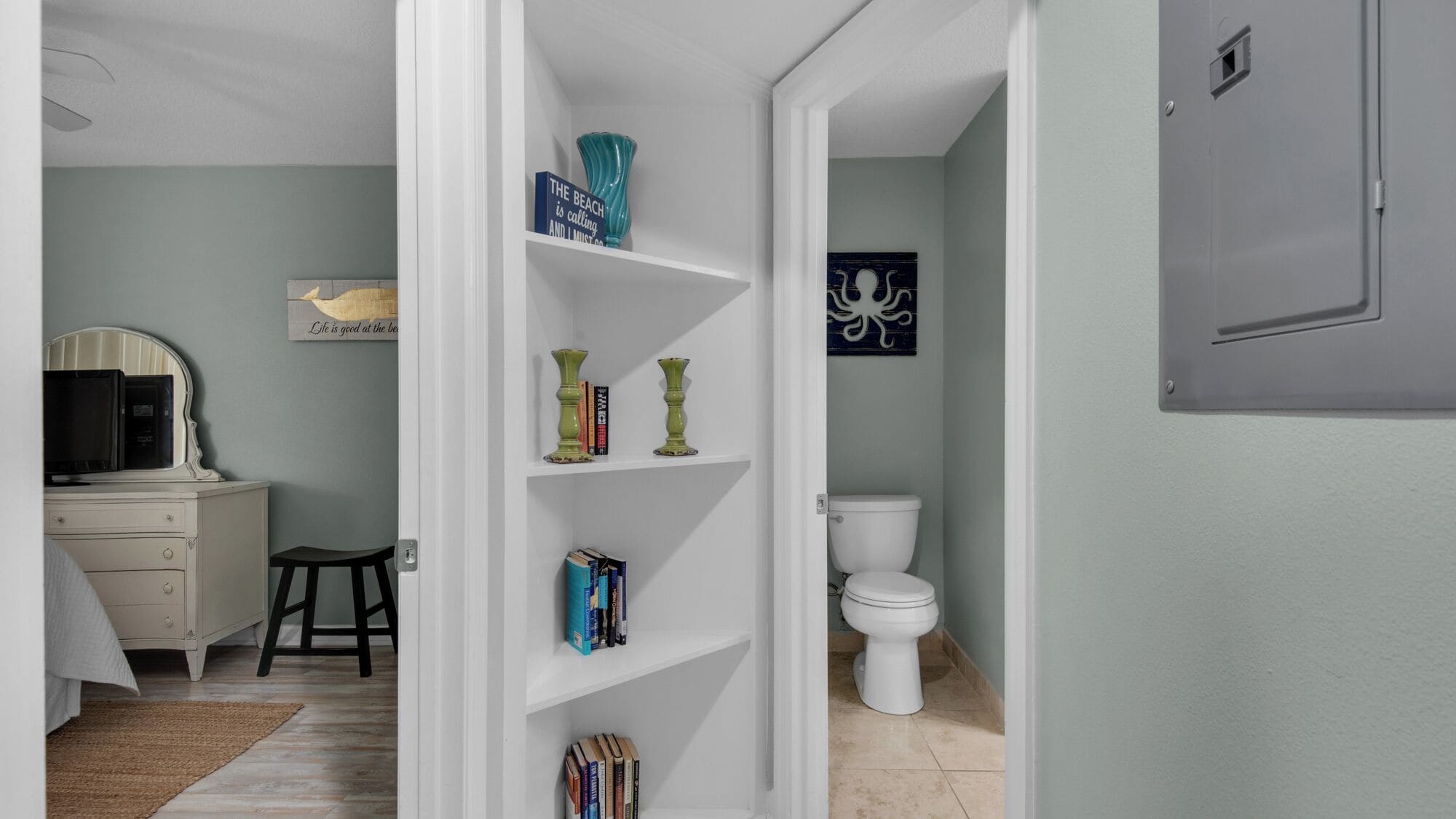 A hallway with a white door leading to a bathroom. There is a white vanity with a mirror and a microwave on top. Shelves with books and decorative items are visible.